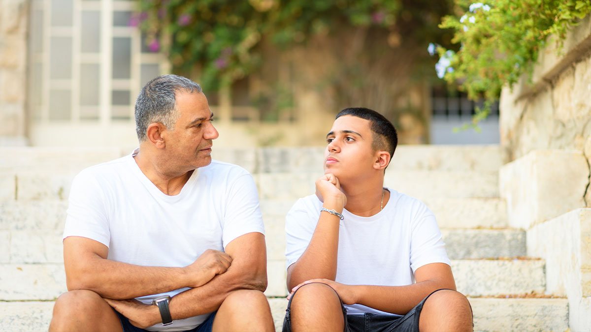 Father talking to teen on stone steps, teen looks like he is thinking about something. 